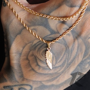 May include: A gold rope chain necklace with a gold angel wing pendant. The necklace is layered on top of another gold rope chain.