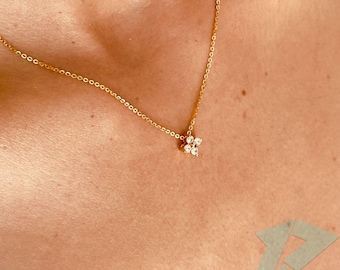 May include: A delicate gold necklace featuring a small, flower-shaped pendant. The pendant is adorned with multiple clear gemstones. The necklace is displayed against a skin tone background, highlighting the jewelry's design and color.