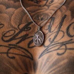 May include: A silver chain necklace with an oval pendant featuring a detailed angel design. The necklace is worn by a person with tattoos.