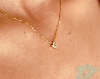 May include: A delicate gold necklace featuring a small, four-petal flower pendant. The pendant is adorned with clear, sparkling gemstones. The necklace is displayed on a person's skin, highlighting its elegant design.