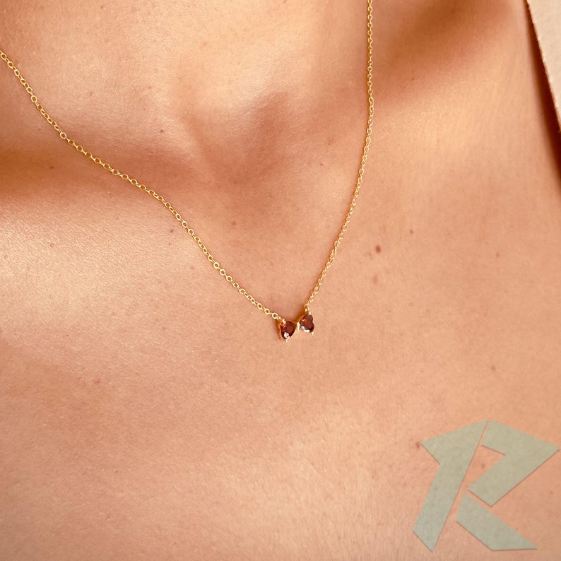 May include: A delicate gold necklace with a fine chain and two heart-shaped, dark red gemstones. The necklace is displayed on a person's chest, highlighting the pendant's design. The gold chain is approximately 45 cm long.
