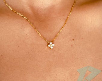 May include: A delicate gold necklace featuring a small, four-petal flower pendant with sparkling white gemstones. The necklace is displayed on a person's chest, showcasing its elegant design. The chain is thin and the pendant is centered.