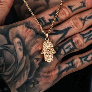 May include: Gold-colored Hamsa hand pendant necklace. The pendant features intricate detailing and is suspended from a matching rope chain. The necklace is a jewelry item.