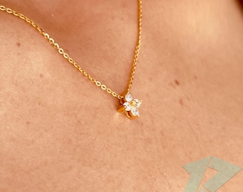 May include: A gold-colored necklace with a delicate chain and a flower-shaped pendant. The pendant features five clear, sparkling gemstones set in a gold-tone metal. The necklace is displayed against a skin-toned background.