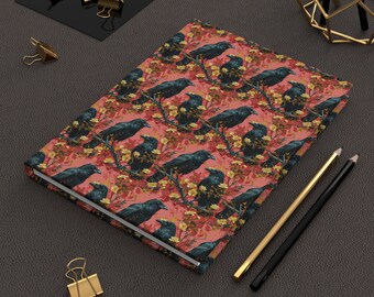 May include: A pink and red notebook with a black crow pattern. The notebook is closed and lying on a dark gray surface.