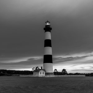 May include: A black and white striped lighthouse stands tall against a cloudy sky. The lighthouse is surrounded by a grassy field and a white fence.