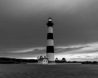 May include: A black and white striped lighthouse stands tall against a cloudy sky. The lighthouse is surrounded by a grassy field and a white fence.