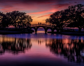 May include: A wooden bridge with two people standing on it, silhouetted against a vibrant sunset sky. The bridge spans a calm body of water, reflecting the colorful sky and the silhouettes of trees on either side.