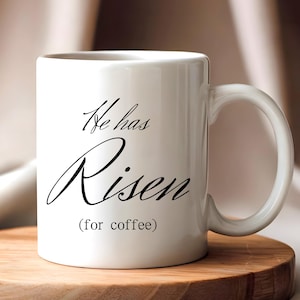 Puede incluir: Taza de café de cerámica blanca con el texto "He has Risen (for coffee)" en escritura negra. La taza tiene un asa curva y está sobre un posavasos de madera. El fondo es de un color neutro suave.