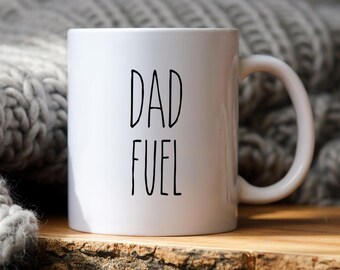 May include: White ceramic mug with the words "DAD FUEL" in black, sans-serif font. The mug has a curved handle and sits on a wooden surface. A gray knitted blanket is in the background.