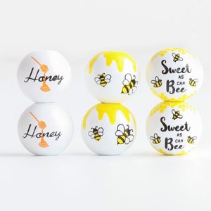 Puede incluir: Seis adornos redondos de madera pintados en amarillo y blanco. Dos adornos dicen "Honey" con un diseño de cucharón de miel. Cuatro adornos dicen "Sweet as can Bee" con un diseño de abeja.
