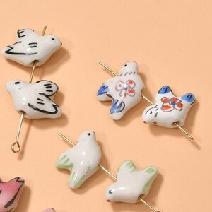 May include: Assortment of ceramic bird charms with gold-colored wire loops. The birds are painted in various colors, including white, blue, pink, and green. Some birds have floral designs.
