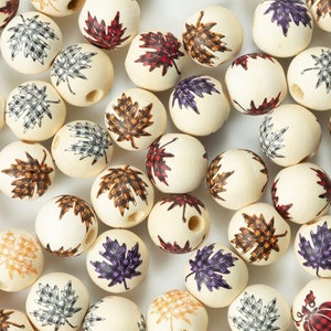 May include: A collection of wooden beads with a variety of autumn leaf designs. The beads are cream coloured with brown, red, purple, and black leaf patterns.