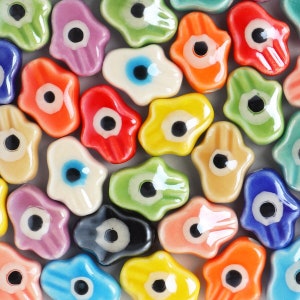 May include: A collection of colourful ceramic hamsa hand charms with an evil eye design. The charms are in various colours, including blue, green, red, yellow, and purple.