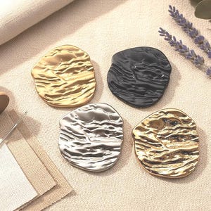 Könnte beinhalten: Vier unregelmäßig geformte, metallische Untersetzer in Gold, Schwarz, Silber und Bronze. Die Untersetzer haben ein strukturiertes, organisches Design. Eine Spule weißen Fadens, eine Nadel und Stoff sind ebenfalls vorhanden.