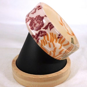 May include: A white bangle bracelet with a floral pattern in shades of orange, brown, and green. The bracelet is displayed on a wooden stand.