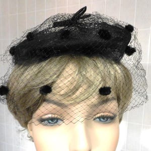 May include: A black pillbox hat with a net veil. The hat features a small bow and is adorned with several black pom-poms. The net veil drapes over the face, creating a vintage and elegant look.