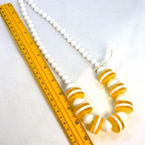 May include: A white beaded necklace with alternating sections of white and yellow-striped beads. A yellow ruler is placed next to the necklace. The necklace is a vintage accessory.