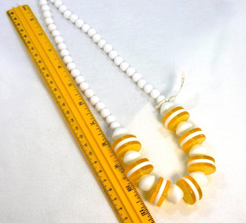 May include: A white beaded necklace with alternating sections of white and yellow-striped beads. A yellow ruler is placed next to the necklace. The necklace is a vintage accessory.