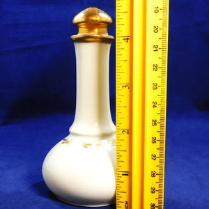 May include: A white ceramic perfume bottle with a gold-colored stopper. The bottle is approximately 5 inches tall. Delicate gold floral accents adorn the lower portion of the bottle. The background is a solid blue color.