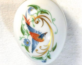 May include: White oval porcelain trinket box with a painted bird and floral design. The bird has blue wings, orange chest, and a red tail. Gold accents and blue flowers decorate the box.