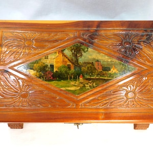 May include: A rectangular wooden box with a hinged lid. The box is made of a light brown wood with a glossy finish. The lid features carved floral designs and a central diamond-shaped panel with a painted landscape scene. The box has small feet and a metal clasp.