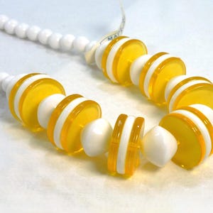May include: A necklace featuring a strand of white, round beads. The focal point is a series of yellow and white disc-shaped beads. The necklace has a tag attached. The design is a mix of bright colors and geometric shapes.