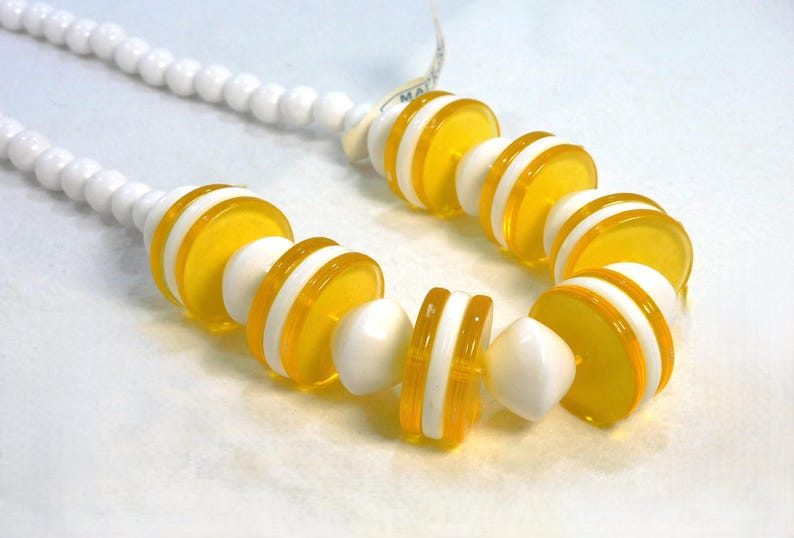 May include: A necklace featuring a strand of white, round beads. The focal point is a series of yellow and white disc-shaped beads. The necklace has a tag attached. The design is a mix of bright colors and geometric shapes.