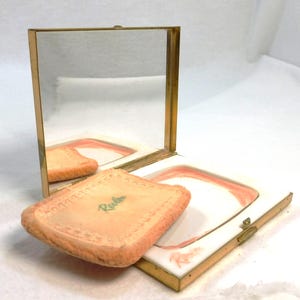 May include: Open vintage compact with a mirror and a powder puff. The compact has a gold-colored frame and a white interior with a pink border. The powder puff is peach-colored with the word "Revlon" printed on it.