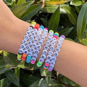 May include: A collection of colorful beaded bracelets spelling out the days of the week. Each bracelet features white letter beads and vibrant, stacked disc beads in various colors. The bracelets are displayed on a wrist against a backdrop of green foliage.
