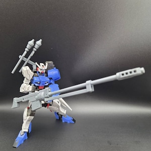 Op de afbeelding: Een wit en blauw robotactiefiguur in een dynamische houding. Het figuur heeft een rood borstdetail, een hoofd met een V-vormige antenne en houdt een lang grijs geweer vast. Twee grijze raketachtige hulpstukken zijn op de rug bevestigd.