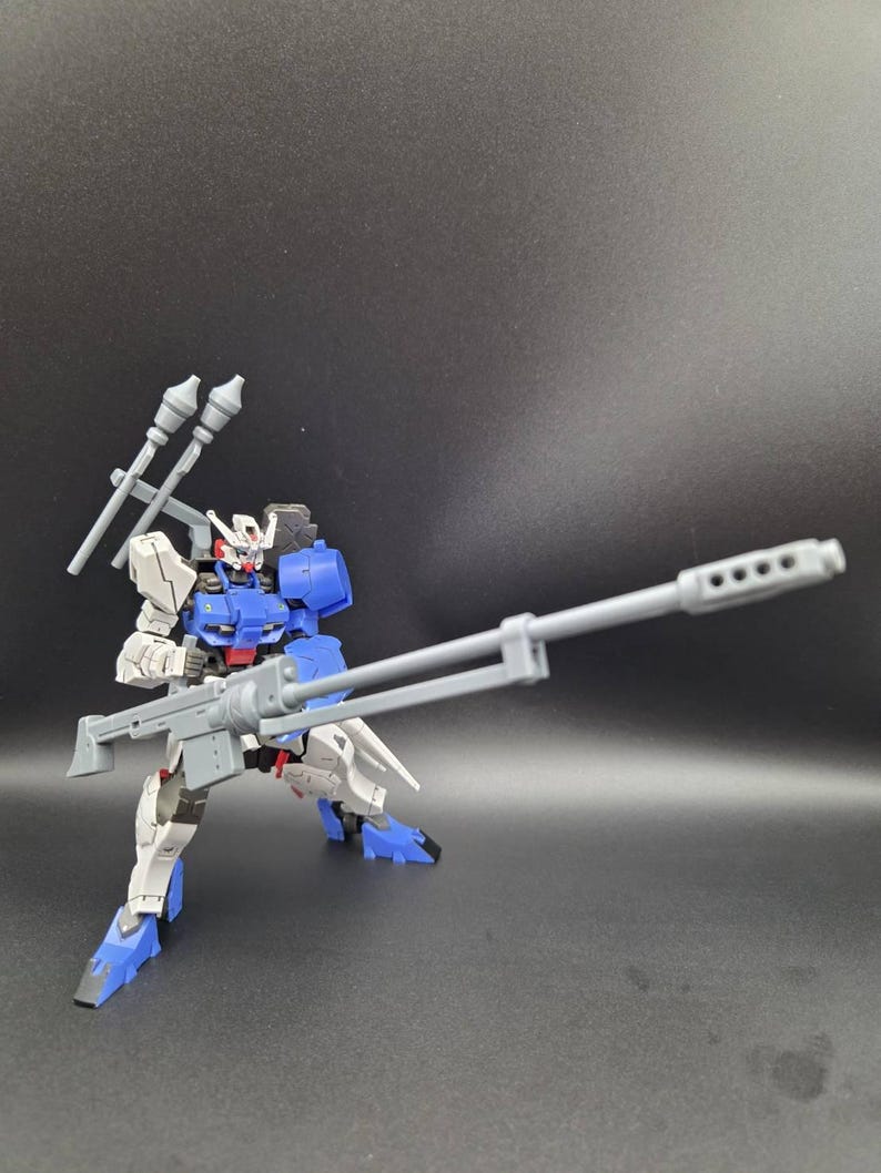 Op de afbeelding: Een wit en blauw robotactiefiguur in een dynamische houding. Het figuur heeft een rood borstdetail, een hoofd met een V-vormige antenne en houdt een lang grijs geweer vast. Twee grijze raketachtige hulpstukken zijn op de rug bevestigd.