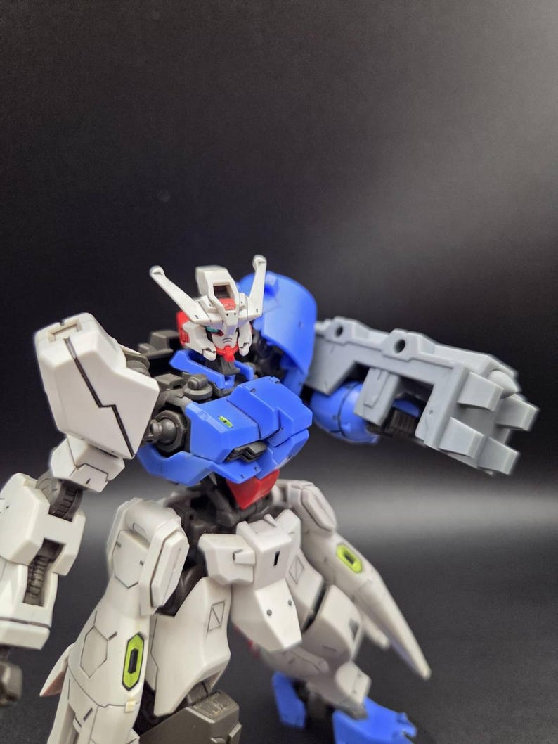 Op de afbeelding: Een plastic robotactiefiguur met een wit, blauw en grijs kleurenschema. Het figuur heeft een gedetailleerd hoofd met een rode kin, een blauwe borstplaat en een grote grijze mechanische armbevestiging. Het ontwerp bevat kleine groene accenten.