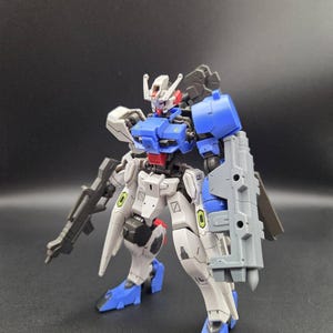 Op de afbeelding: Een beweegbaar robotactiefiguur in een wit, blauw en grijs kleurenschema. Het figuur heeft een gedetailleerd hoofd met een rode kin, een blauw borststuk en een groot grijs schild aan de linkerarm. Het houdt een grijs geweer in de rechterhand.