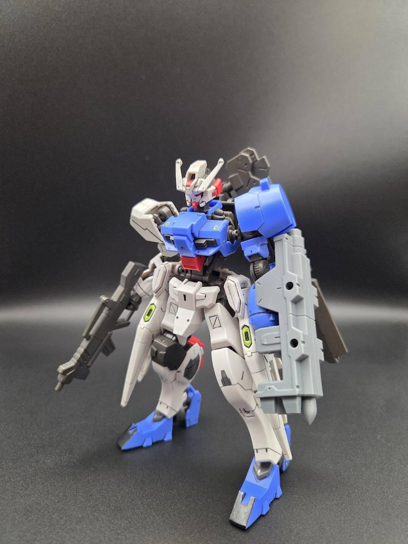 Op de afbeelding: Een beweegbaar robotactiefiguur in een wit, blauw en grijs kleurenschema. Het figuur heeft een gedetailleerd hoofd met een rode kin, een blauw borststuk en een groot grijs schild aan de linkerarm. Het houdt een grijs geweer in de rechterhand.
