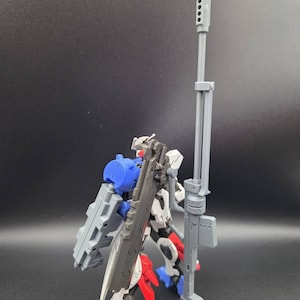 Op de afbeelding: Een plastic robotactiefiguur met een wit, blauw en rood kleurenschema. Het figuur is uitgerust met een groot, hoog grijs geweer en een donkergrijs zwaardachtig wapen op de rug, met gedetailleerde mechanische onderdelen en structuren.