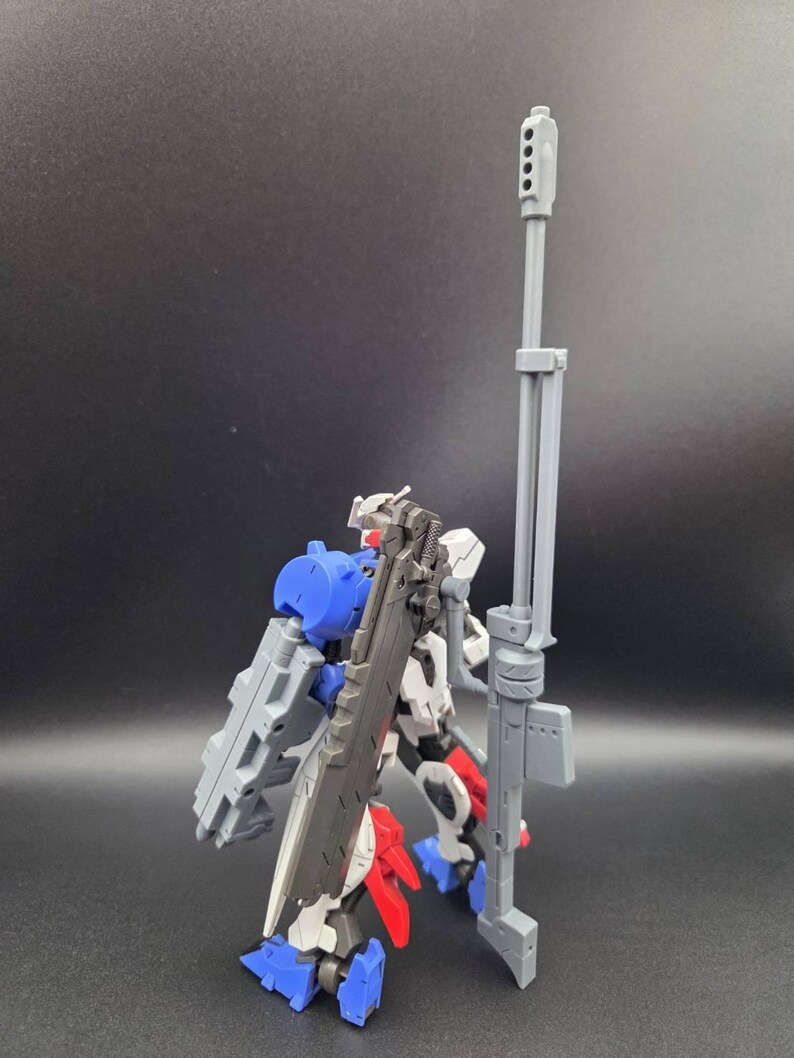 Op de afbeelding: Een plastic robotactiefiguur met een wit, blauw en rood kleurenschema. Het figuur is uitgerust met een groot, hoog grijs geweer en een donkergrijs zwaardachtig wapen op de rug, met gedetailleerde mechanische onderdelen en structuren.