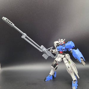 Op de afbeelding: Een beweegbaar robotactiefiguur met een wit, blauw en rood kleurenschema. Het figuur is uitgerust met een groot, grijs geweer met een lange loop in de rechterhand. Het heeft een gedetailleerd hoofd, beweegbare ledematen en blauwe pantserplaten op de schouders, voeten en de linkeronderarm.