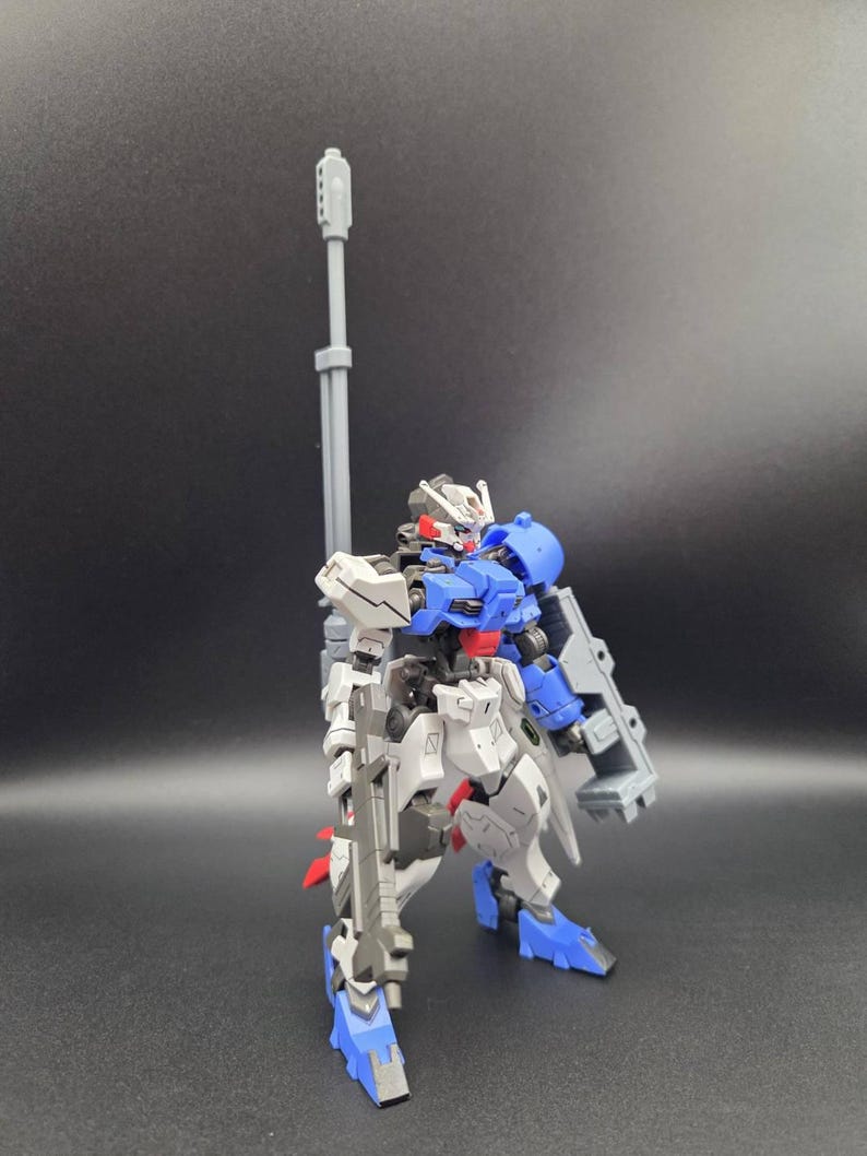 Op de afbeelding: Een beweegbaar robotactiefiguur met een blauw, wit en grijs kleurenschema. Het figuur is uitgerust met een lang grijs kanon op de rug, een grijs geweer in de hand en een groot grijs schild bevestigd aan de linkerarm.