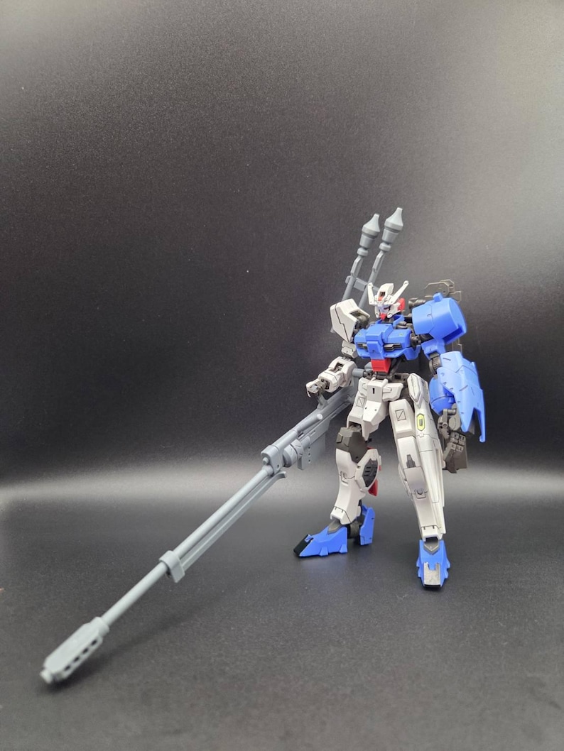 Op de afbeelding: Een wit, blauw en grijs robotactiefiguur dat rechtop staat. Het figuur heeft een gedetailleerd mechanisch ontwerp met een lang grijs geweer in de rechterhand, twee grijze kanonnen op de rug en blauwe schouderpantserplaten.