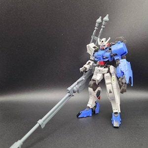 Op de afbeelding: Een plastic modelrobotfiguur met een wit, blauw en grijs kleurenschema. Het figuur heeft een humano&iuml;de structuur met beweegbare ledematen, een hoofd met een V-vormige antenne en is uitgerust met een lang grijs geweer en twee verticale grijze kanonnen.