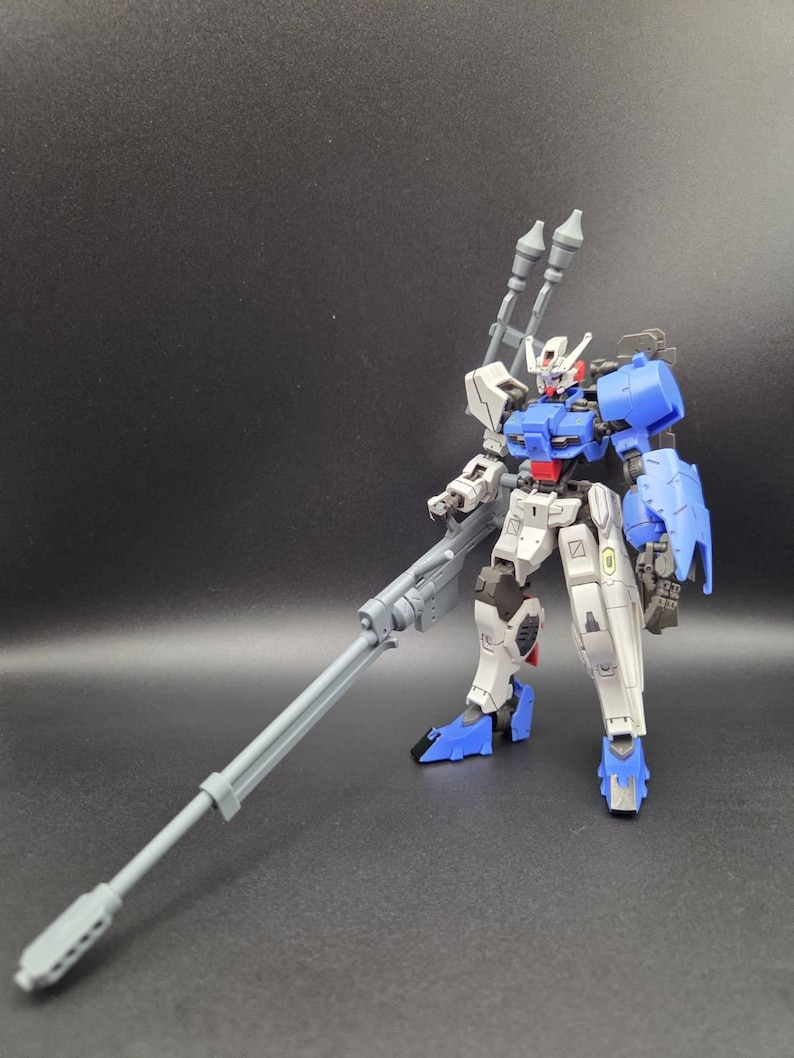 Op de afbeelding: Een plastic modelrobotfiguur met een wit, blauw en grijs kleurenschema. Het figuur heeft een humano&iuml;de structuur met beweegbare ledematen, een hoofd met een V-vormige antenne en is uitgerust met een lang grijs geweer en twee verticale grijze kanonnen.