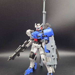Op de afbeelding: Een verstelbaar robotactiefiguur met een wit, blauw en grijs kleurenschema. Het figuur is uitgerust met een lang grijs kanon op de rug, een geweer in de hand en een groot grijs schild bevestigd aan de linkerarm. Het heeft een gedetailleerd hoofd.