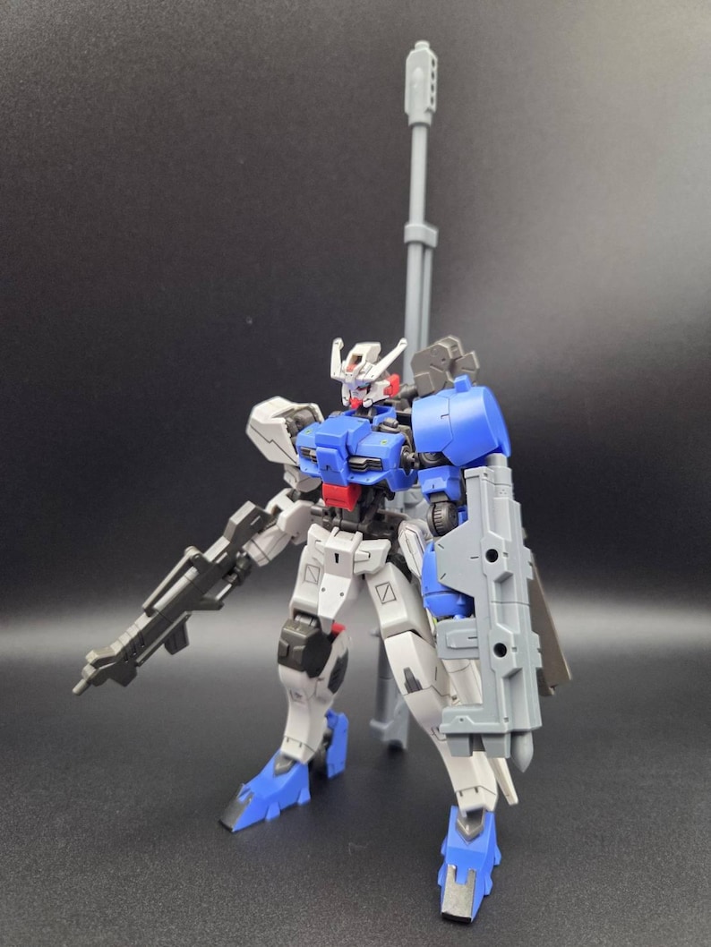 Op de afbeelding: Een verstelbaar robotactiefiguur met een wit, blauw en grijs kleurenschema. Het figuur is uitgerust met een lang grijs kanon op de rug, een geweer in de hand en een groot grijs schild bevestigd aan de linkerarm. Het heeft een gedetailleerd hoofd.