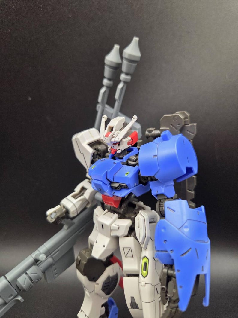Op de afbeelding: Een plastic robotfiguur met een kleurenschema van wit, blauw en grijs. Het figuur heeft een gedetailleerd hoofd met rode accenten, een blauw borststuk en een groot grijs wapen op de rug. Het staat met beweegbare ledematen.