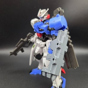 Op de afbeelding: Een plastic robotfiguur met een wit, blauw en grijs kleurenschema. Het figuur heeft een hoofd met een V-vormige antenne, een blauw borststuk, een groot grijs schild bevestigd aan de linkerarm en een grijs geweer in de hand. Het staat in een pose.