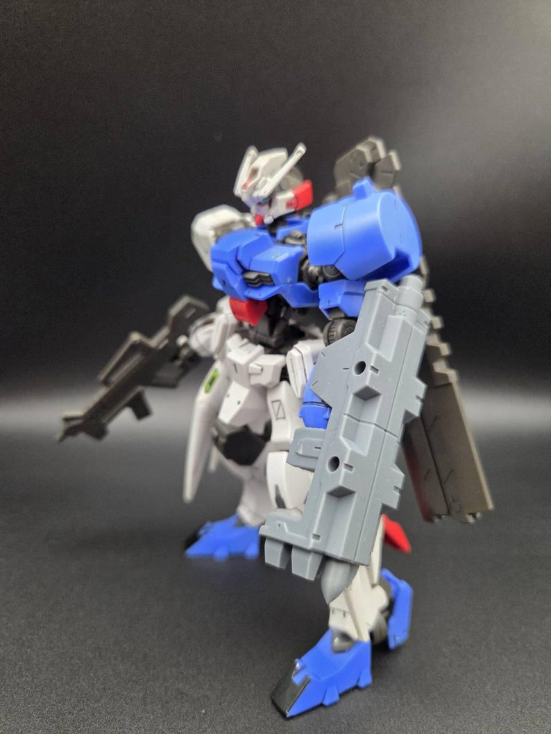 Op de afbeelding: Een plastic robotfiguur met een wit, blauw en grijs kleurenschema. Het figuur heeft een hoofd met een V-vormige antenne, een blauw borststuk, een groot grijs schild bevestigd aan de linkerarm en een grijs geweer in de hand. Het staat in een pose.