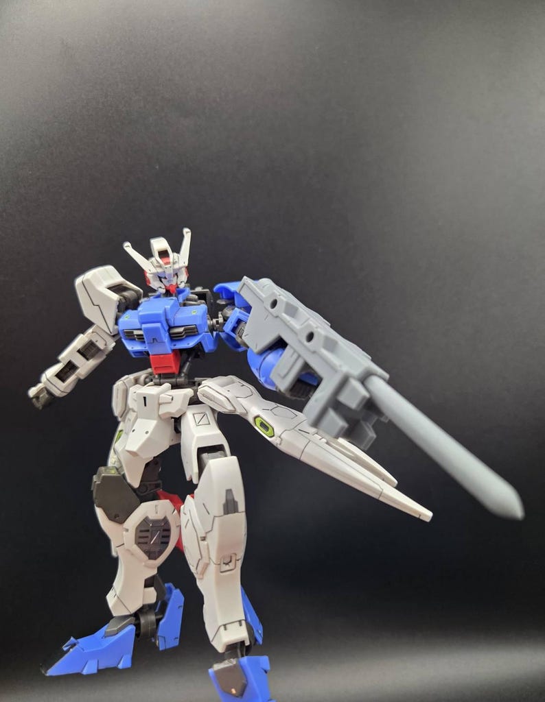 Op de afbeelding: Een beweegbaar robotactiefiguur met een wit, blauw en rood kleurenschema. Het figuur heeft een gedetailleerd mechanisch ontwerp met beweegbare ledematen, een hoofd met een V-vormige kam en een groot grijs wapen aan de linkerarm.