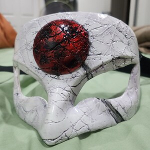 Puede incluir: Una máscara blanca con textura agrietada y un gran círculo rojo texturizado en el centro. La máscara tiene aberturas para ojos y nariz, probablemente para disfraz o decoración.