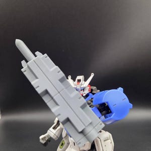 Op de afbeelding: Een wit, blauw en grijs gearticuleerd robotactiefiguur dat een groot, rechthoekig grijs wapen vasthoudt. Het figuur heeft een gedetailleerd hoofd met rode accenten, een blauw schouderstuk en mechanische beencomponenten met groene accenten.